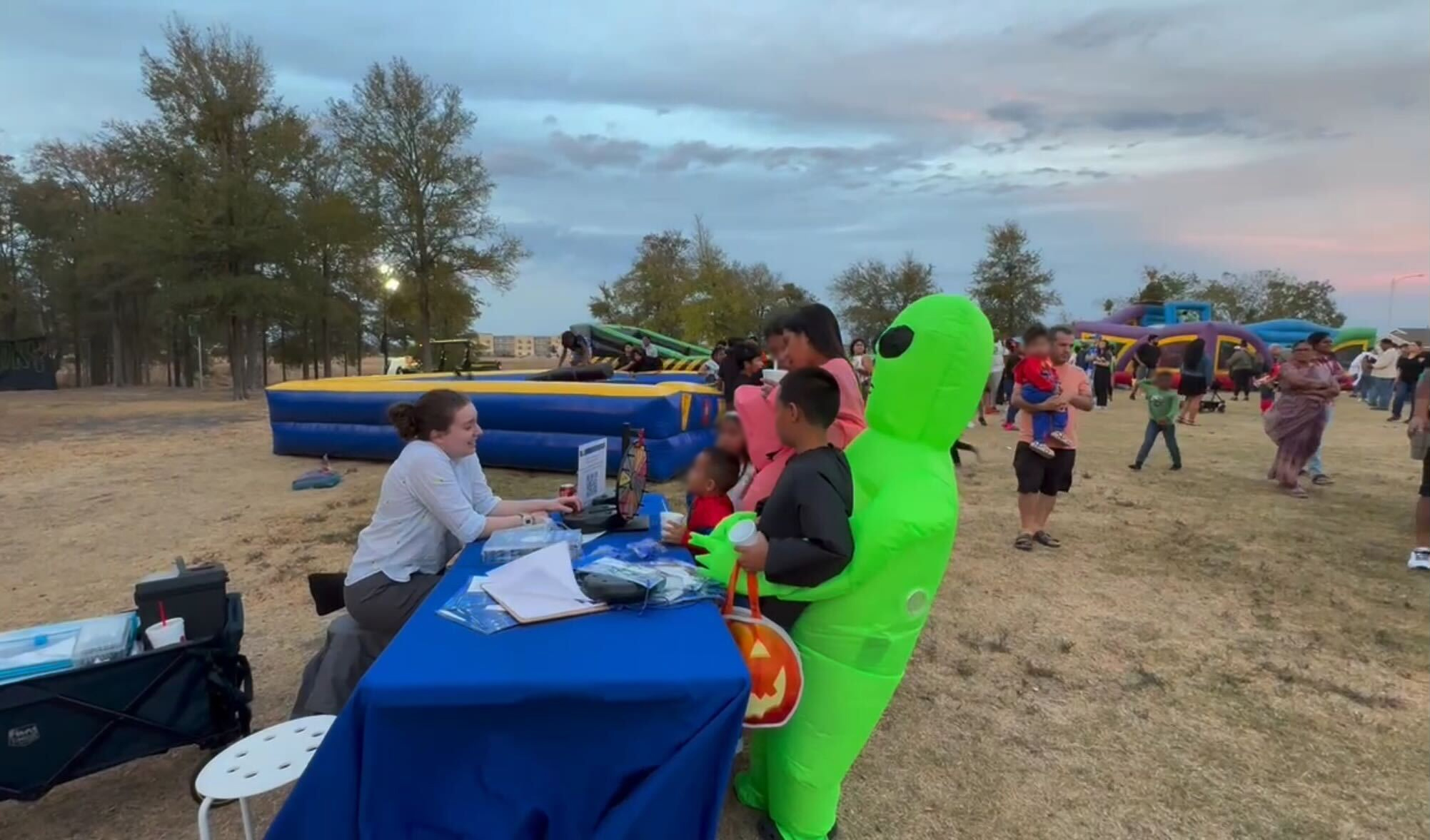 A person in a bright green alien costume and others in colorful outfits speak with a staff member at an outdoor booth covered with a blue tablecloth during a community event.